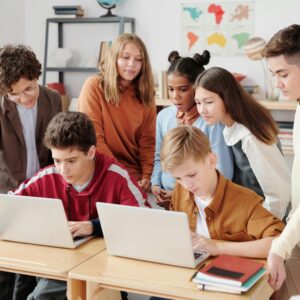 A group of diverse students collaborating on laptops in a classroom setting, guided by a teacher.